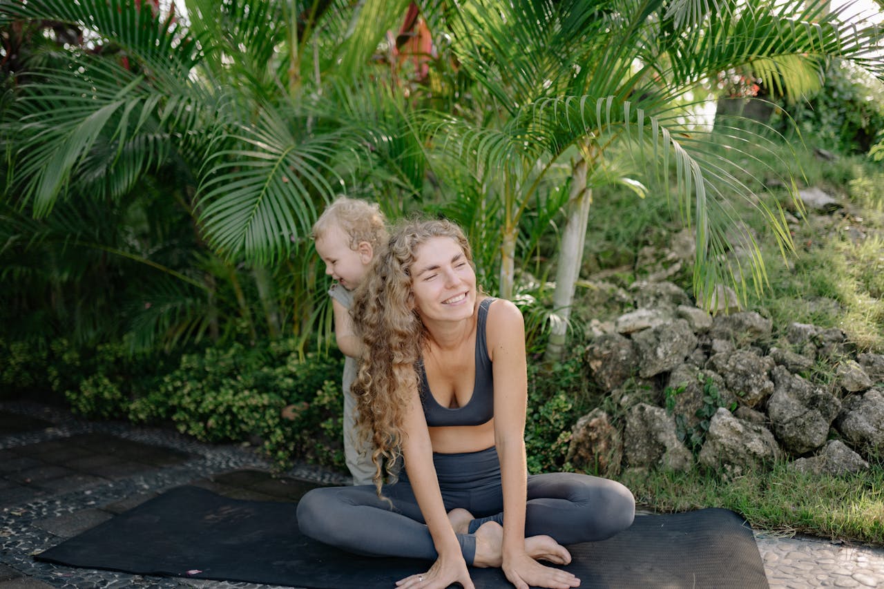 Cheerful smiling sportswoman sitting in lotus pose with adorable little son on yoga mat in green tropical garden